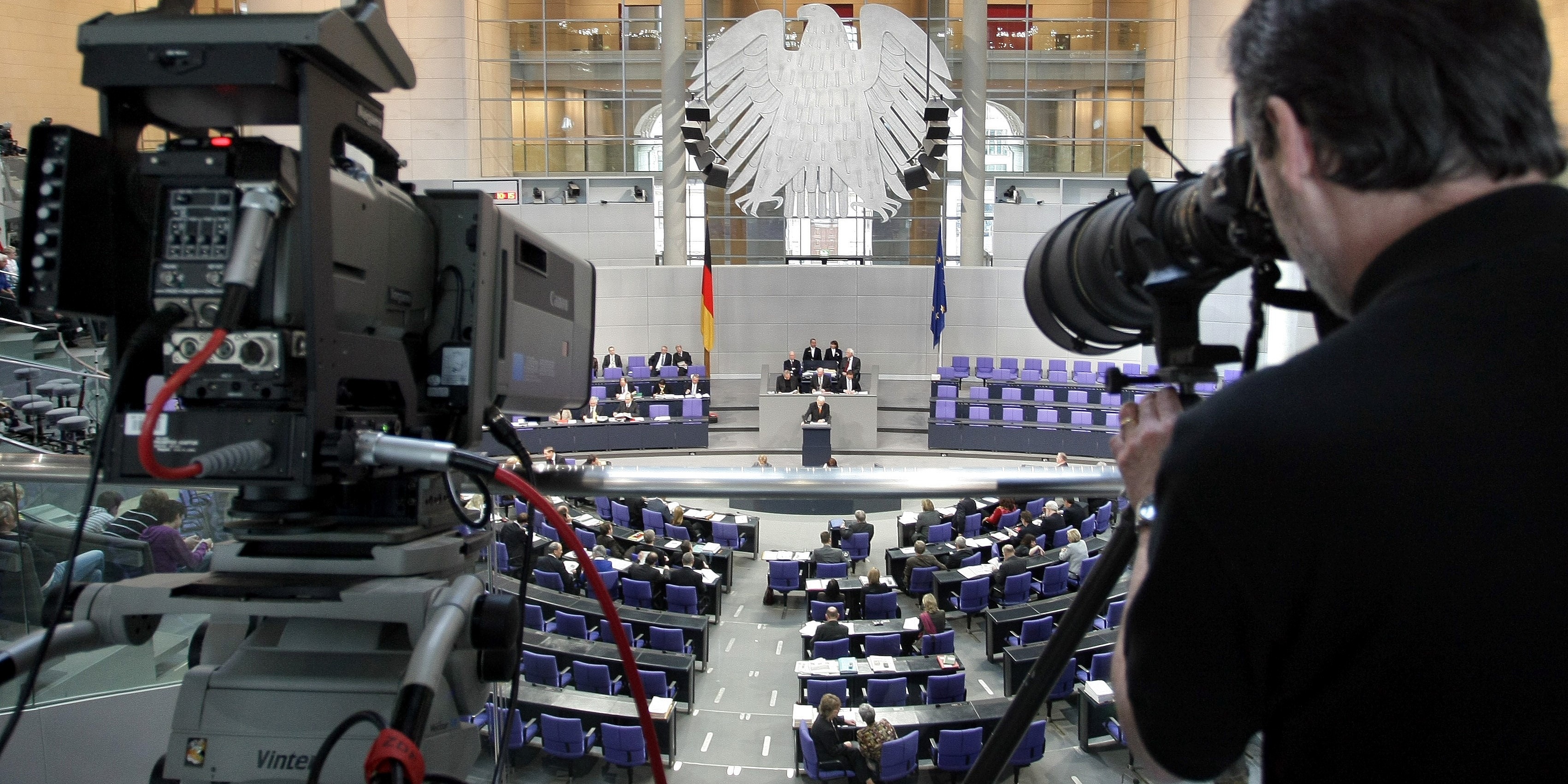 Von der Pressetribüne des Plenarsaals im Deutschen Bundestag fotografiert ein Journalist in Richtung des Redner_innenpultes. Unten im Saal sind die Abgeordneten, vor allem aber das Podium der Sitzungsleitung und die Sitzreihen der Bundesregierung zu sehen. 