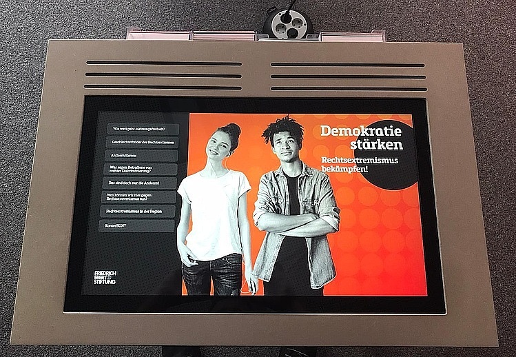 Foto eines Bildschirms. Auf dem Bildschirm eine Präsentationsfolie, auf der ein schwarz-weiß Foto von zwei jungen Menschen abgebildet ist. Auf der Folie weißer Text: "Demokratie stärken - Rechtsextremismus bekämpfen".