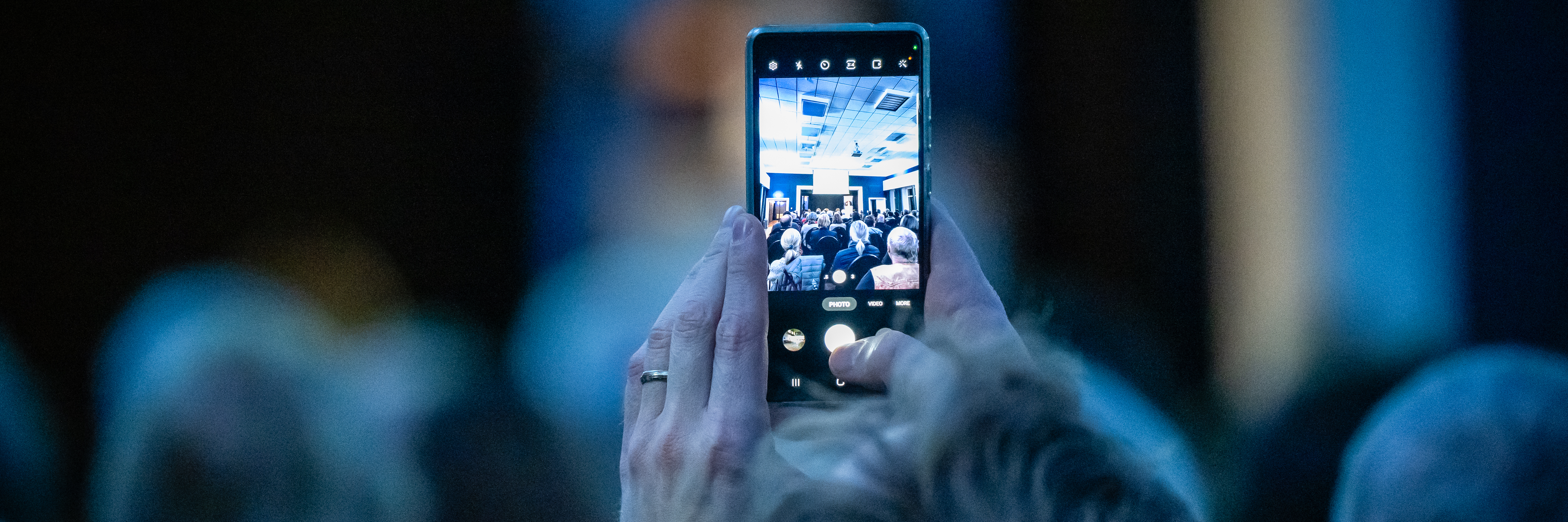 Das Bild zeigt einen Veranstaltungsraum mit Publikum. Eine Person hält ihr Handy in die Höhe und fotografiert die Bühne. 