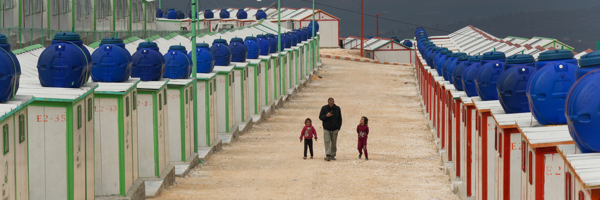 Blick auf einen Weg in einem Flüchtlingslager in Syrien zwischen nebeneinander aufgereihten identisch aussehenden Unterkünften. Auf dem Weg laufen ein Mann mit zwei kleinen Kindern rechts und links von ihm. 