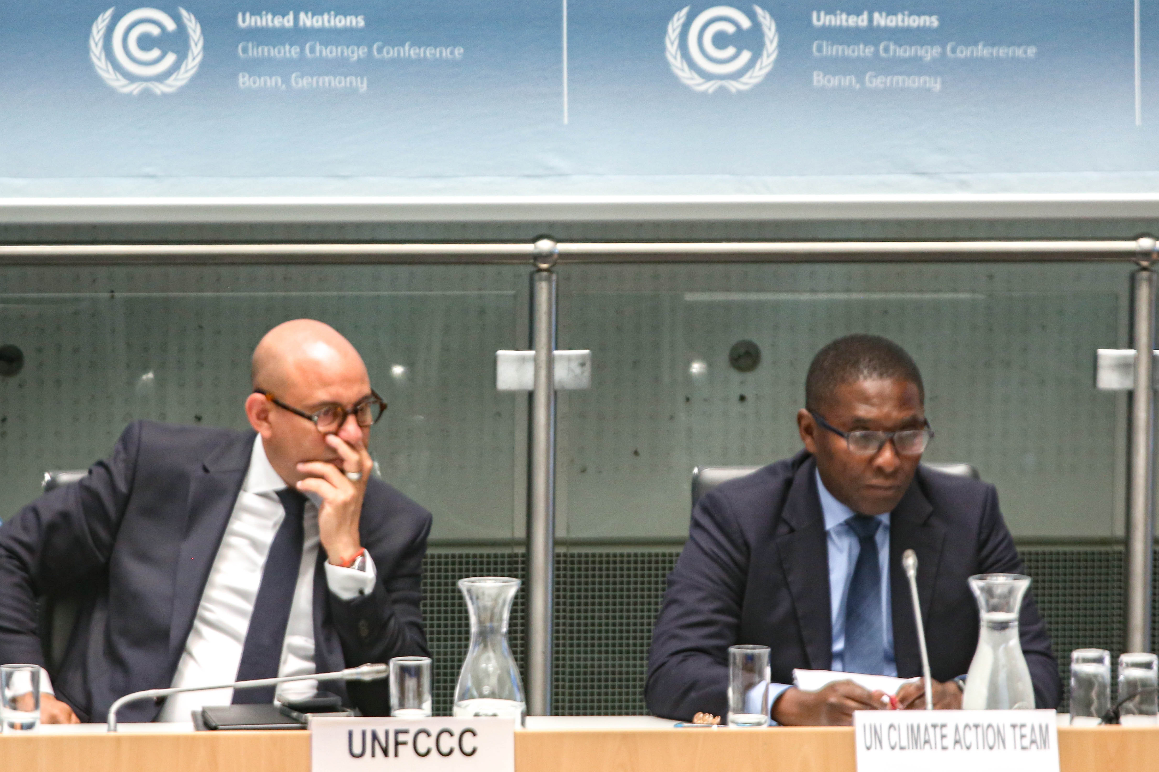 Simon Stiell, Generalsekretär des United Nations Framework on Climate Change sitzt auf dem Podium, hört einem Bericht zu indem er mit der rechten Hand seinen Mund verbirgt. Die Geste wirkt zuückhaltend. Zu seiner Linken sitzt ein weiterer Mann vom UN Climate Team 