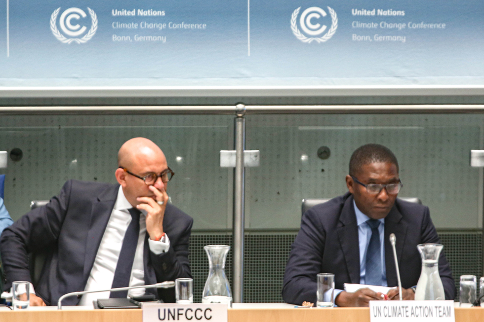 Simon Stiell, Generalsekretär des United Nations Framework on Climate Change sitzt auf dem Podium, hört einem Bericht zu indem er mit der rechten Hand seinen Mund verbirgt. Die Geste wirkt zuückhaltend. Zu seiner Linken sitzt ein weiterer Mann vom UN Climate Team 