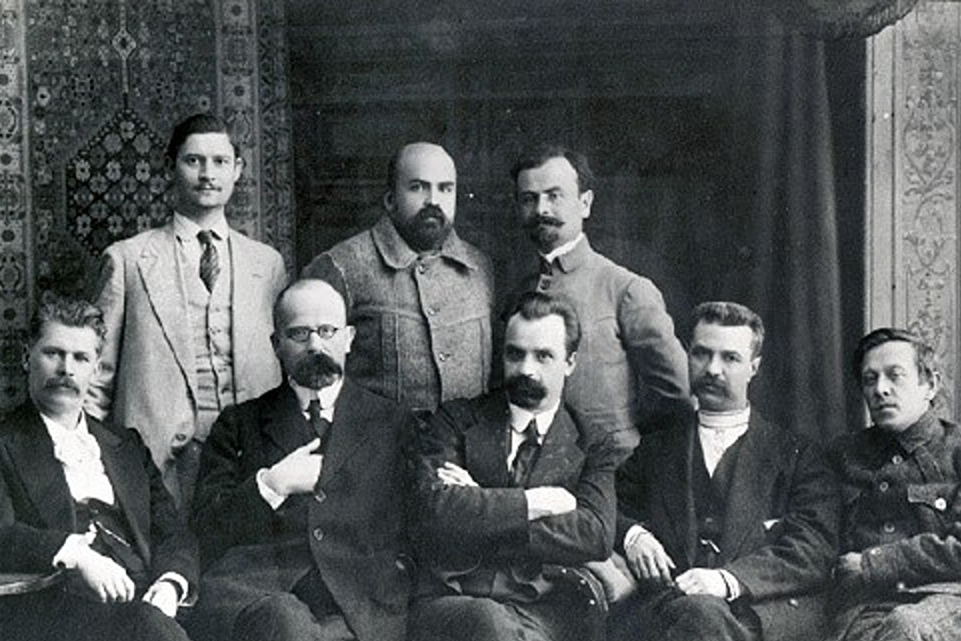 Schwarz-Weiß. Fünf Männer in Anzügen sitzen auf einer Bank. Hinter ihnen stehen drei weitere Männer in Anzügen.