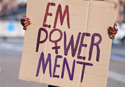 Person hält ein handgemachtes Plakat mit der Aufschrift 'EMPOWERMENT' in lila und roter Farbe, wobei das 'M' und 'W' besonders hervorgehoben sind. Der Hintergrund ist unscharf mit städtischer Kulisse.