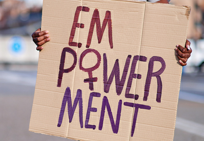 Person hält ein handgemachtes Plakat mit der Aufschrift 'EMPOWERMENT' in lila und roter Farbe, wobei das 'M' und 'W' besonders hervorgehoben sind. Der Hintergrund ist unscharf mit städtischer Kulisse.