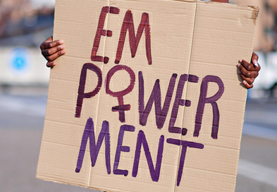 Person hält ein handgemachtes Plakat mit der Aufschrift 'EMPOWERMENT' in lila und roter Farbe, wobei das 'M' und 'W' besonders hervorgehoben sind. Der Hintergrund ist unscharf mit städtischer Kulisse.