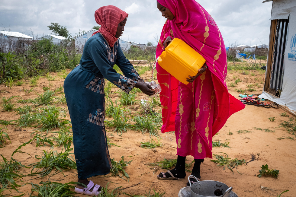 Zwei Frauen in Gewändern reinigen Kaffeetassen mit Hilfe eines Wasserkansiters, im Hintergrund erkennt man ein Camp für Geflüchtete.