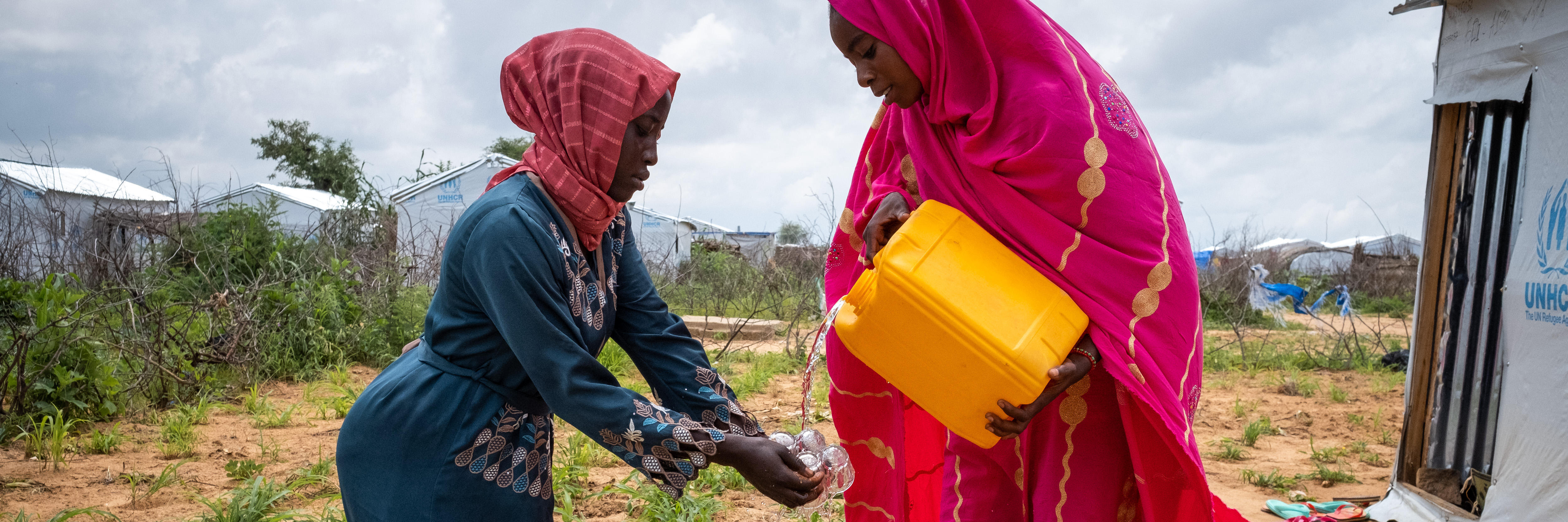 Zwei Frauen in Gewändern reinigen Kaffeetassen mit Hilfe eines Wasserkansiters, im Hintergrund erkennt man ein Camp für Geflüchtete.