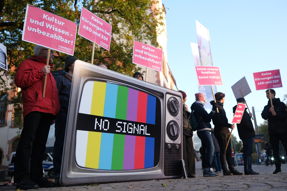 Demonstration gegen Kürzungen in der Medien und Kultur Branche, Menschen mit Schildern, im Vordergrund ein großer Fernseher mit einem bunten "no signal" Testbild.