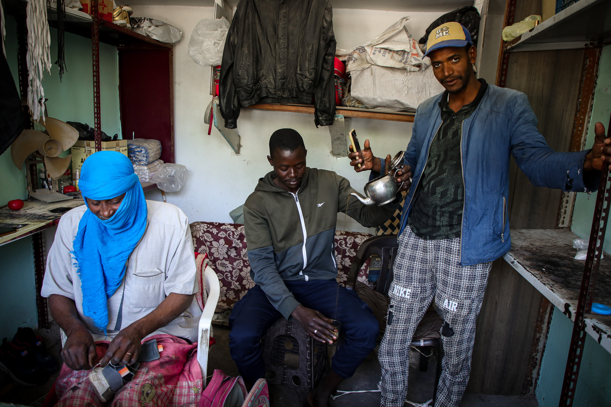 Three migrant workers in Tripoli, Libya, at a sales stall. A man with a bright blue turban is working on a shoe, one is preparing tea and another is looking at the camera with a mobile phone in his hand.
